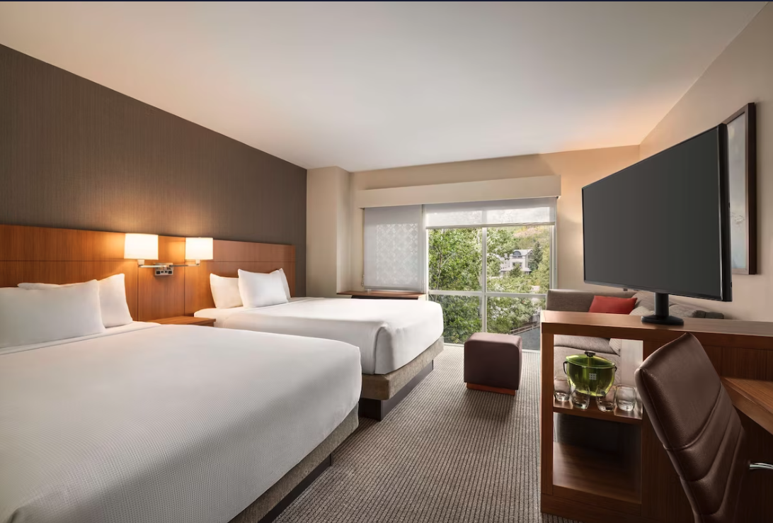 Modern hotel room at Hyatt Place Keystone with two beds, a large TV, a sitting area, and a window overlooking greenery.